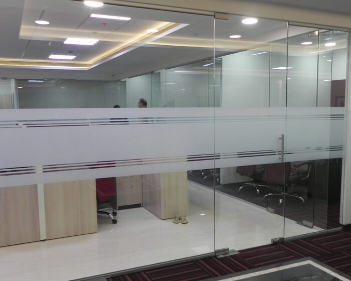 Glass partition cabin view with open workstation desks and maroon ergonomic office chairs